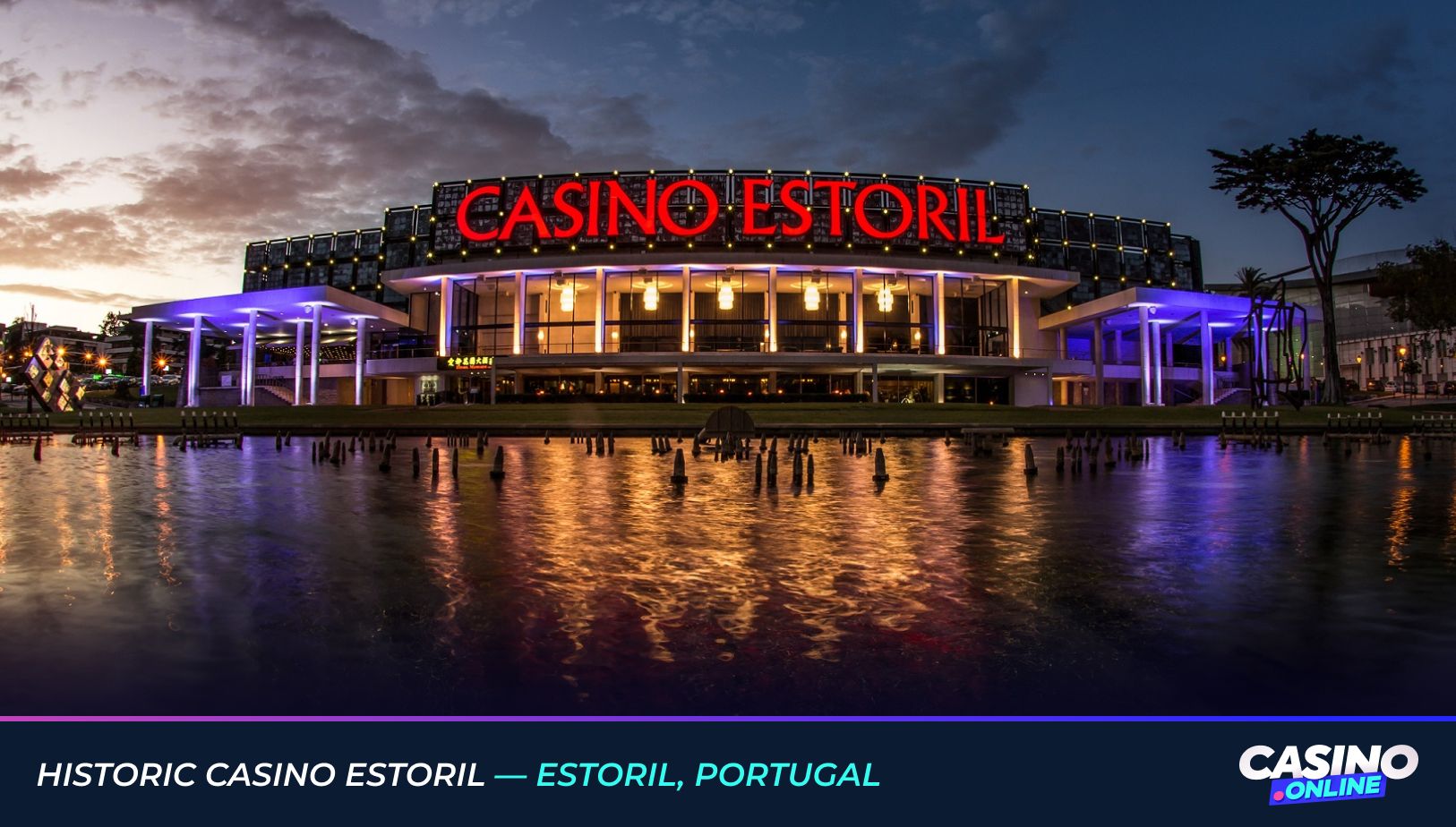 Evening view of Historic Casino Estoril in Estoril, Portugal, with illuminated red signage reflected in the water.